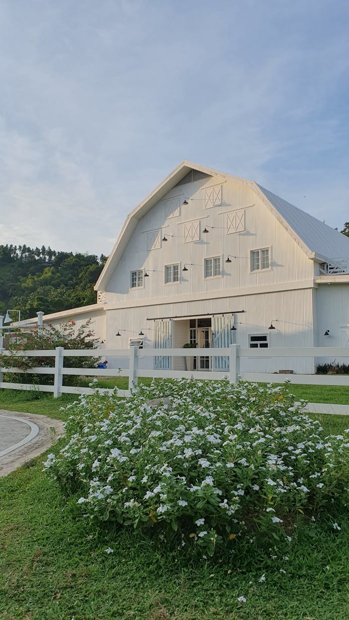 about-us A picturesque white barn surrounded by greenery and flowers on a sunny day.