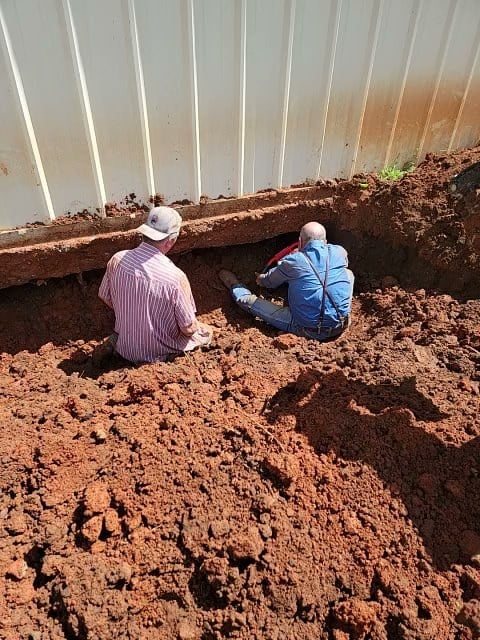 Two workers in a red clay trench beneath a barndominium slab edge running new plumbing