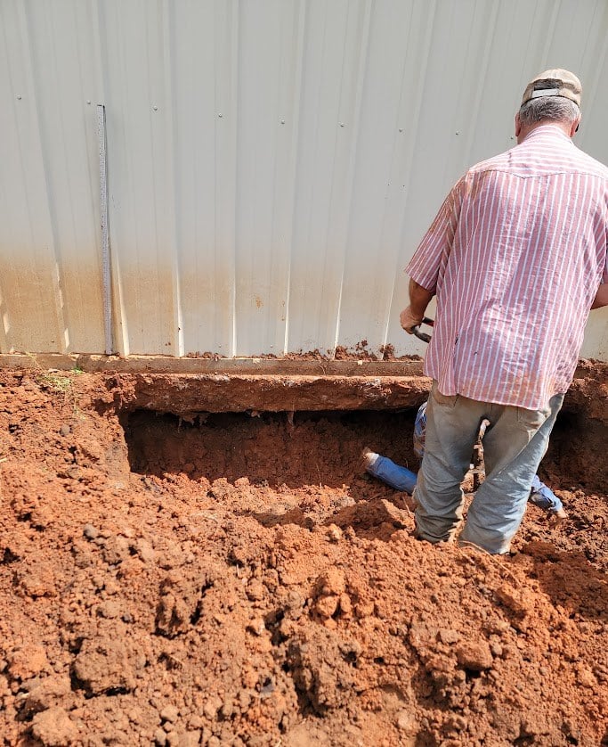 Worker digging red clay beside a metal barndominium wall, exposing the edge of the concrete slab