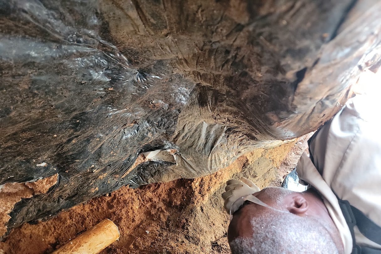 Plumber lying in a red clay trench beneath a concrete slab and its black vapor barrier, preparing to install plumbing
