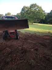 Orange tractor with a front loader backfilling a dirt trench beside a barndominium